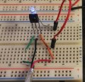 A close-up image of an electronic breadboard setup showing a wired IR receiver and a lit blue LED. The receiver has three legs connected with jumper wires to the extended cable to the amp.